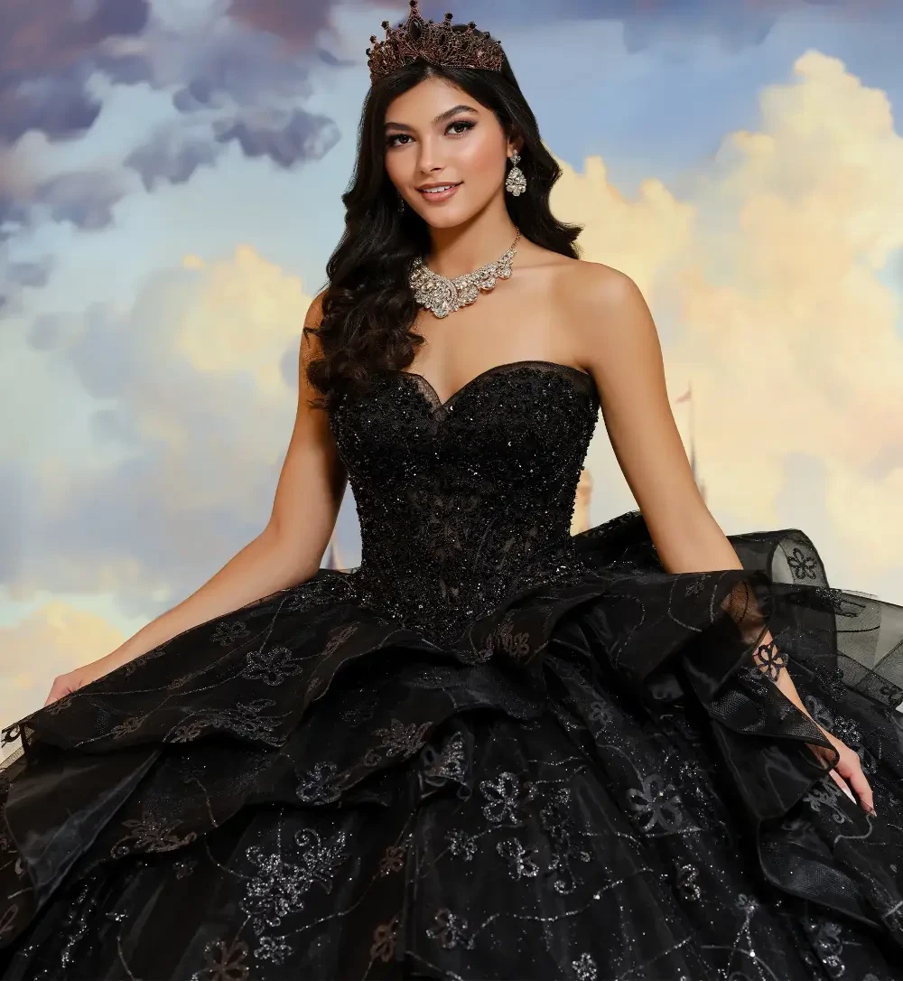 A woman wearing a strapless black gown with layers of ruffles and floral designs, standing in front of a castle backdrop. She has long dark hair and is adorned with a crown and necklace.