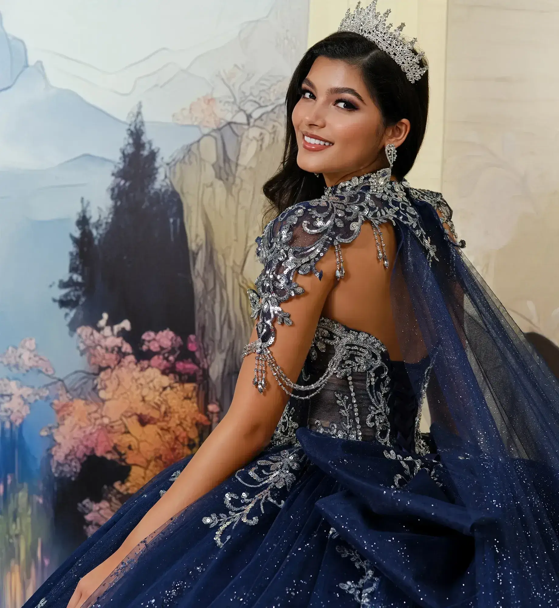 Mujer sonriente con un brillante vestido azul marino con intrincados detalles plateados y una tiara, de pie ante un colorido tel&oacute;n de fondo de tem&aacute;tica natural.  &nbsp;