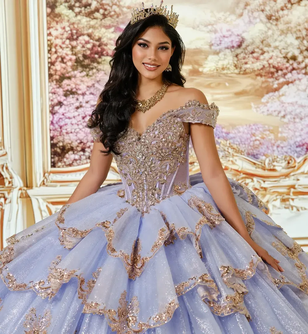 Una joven con un lujoso vestido de baile de hombros descubiertos, con capas de volantes y brillos, de pie en un entorno ornamentado con fondos florales.